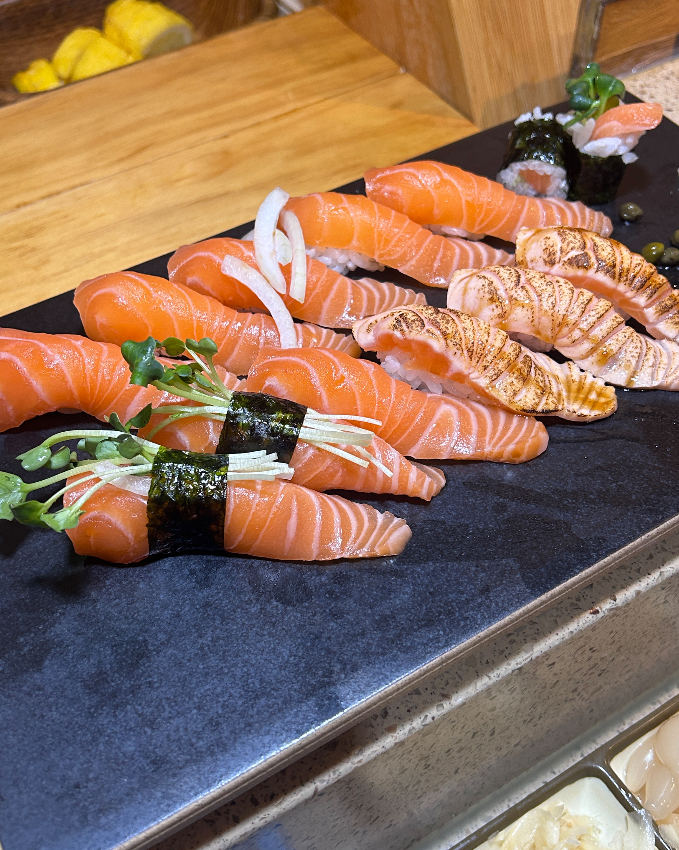 다양한 종류의 초밥이 나무 접시에 보기 좋게 담겨 있고, 곁들임 채소와 소스가 함께 나온다.