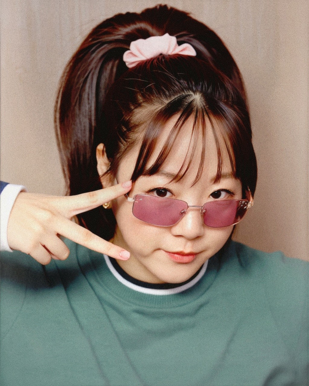 핑크색 틴트 선글라스를 쓴 여성이 브이 포즈를 취하고 있다. 그녀는 묶은 머리에 분홍색 머리끈을 하고 있다.