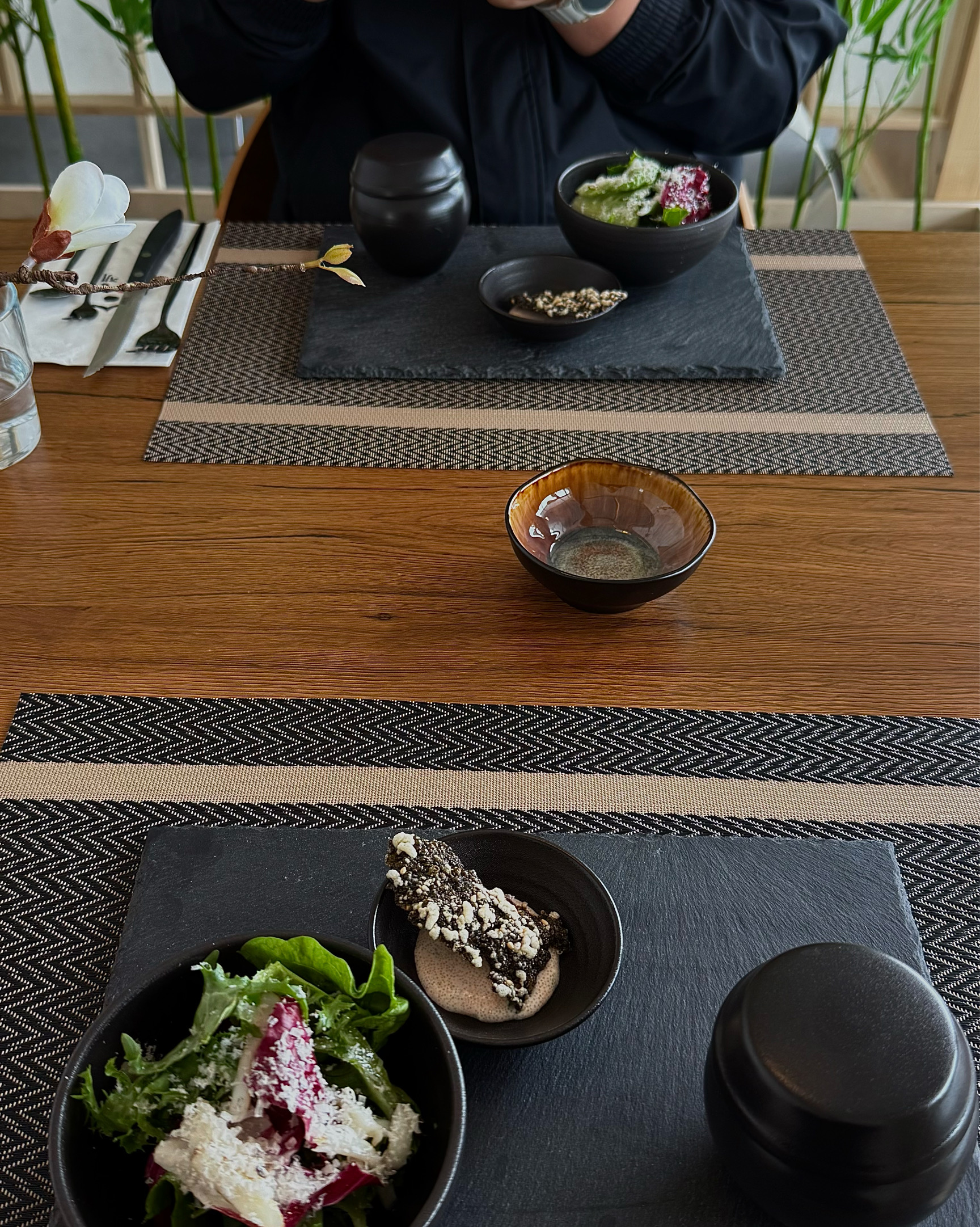 테이블 위에 놓인 검은색 접시에 샐러드와 함께 디저트로 보이는 음식이 담겨 있다. 그 뒤로는 짙은 색의 뚜껑 달린 그릇과 찻잔이 보인다.