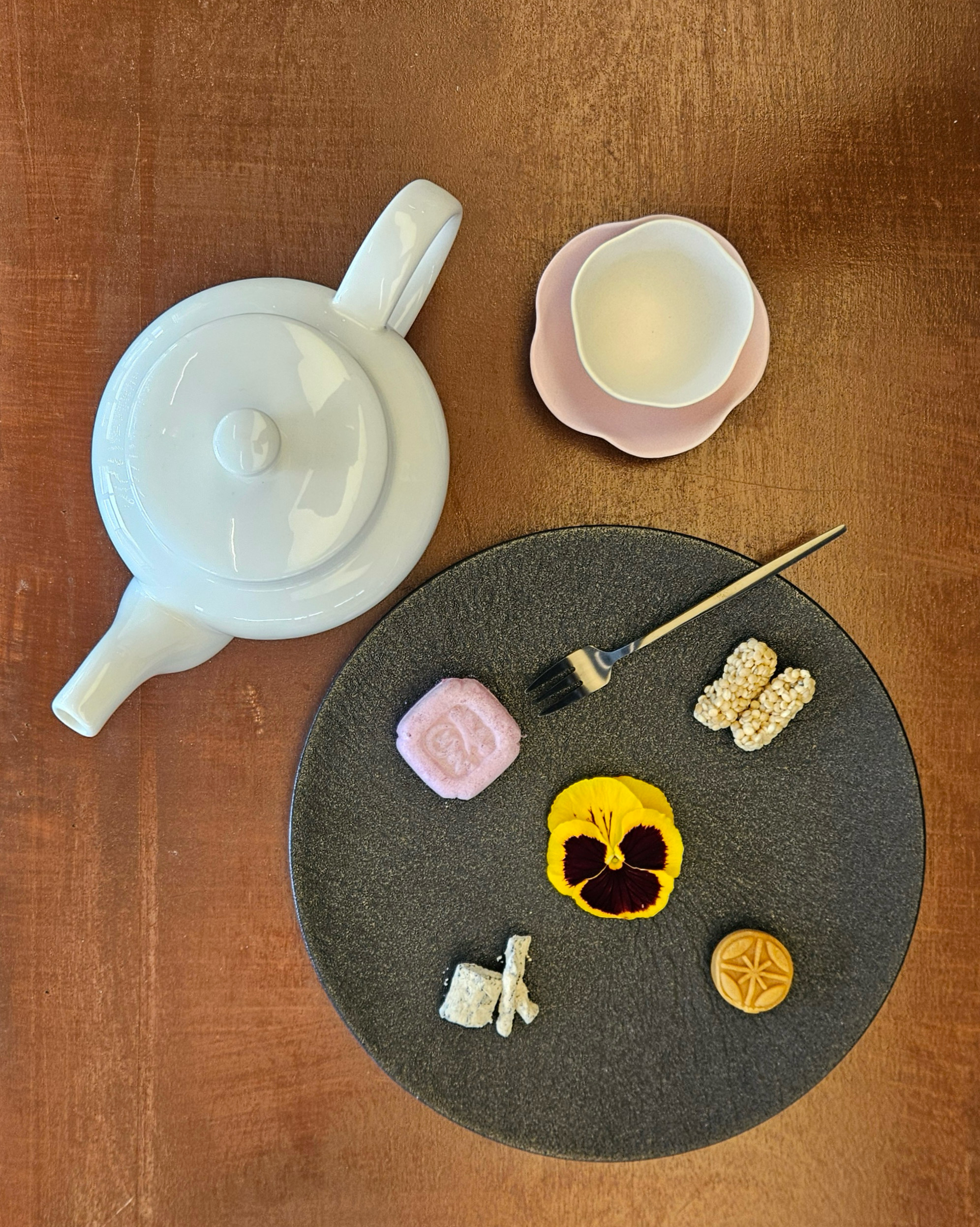 하얀색 티팟과 핑크색 찻잔 세트, 그리고 검은색 접시 위에 다양한 모양의 다과와 꽃잎이 놓여 있다.